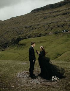 a man and woman dressed in black standing next to each other on top of a lush green hillside