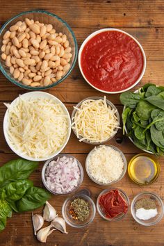 the ingredients to make this pasta dish are laid out on a wooden surface, including spinach leaves, sunflower seeds, red sauce and cheese