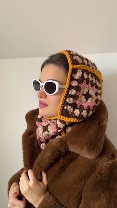 a woman wearing sunglasses and a brown jacket with flowers on it, standing in front of a white wall