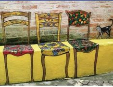 three wooden chairs sitting on top of a yellow table covered in colorful cloths next to a black and white cat