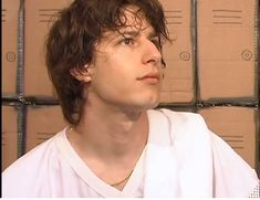 a young man with curly hair wearing a white shirt in front of stacks of boxes