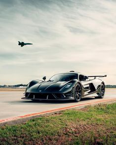 a black sports car driving down the road with an airplane in the sky above it