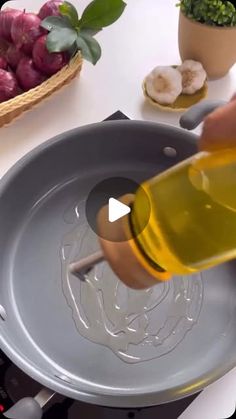 a person pouring oil into a pan on top of a stove with vegetables in the background