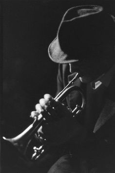 black and white photograph of a man playing the trumpet in front of his face, wearing a fedora