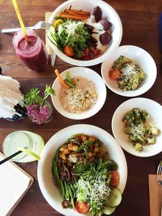 four white bowls filled with different types of food on top of a wooden table next to drinks and utensils