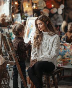 a woman sitting next to a little boy in front of an easel and painting