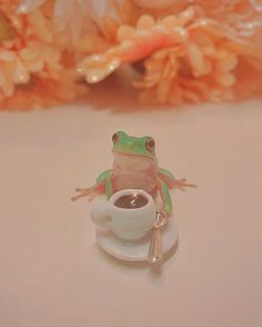 a tiny frog sitting on top of a white saucer next to a coffee cup