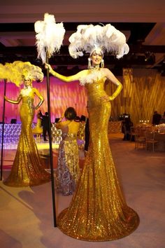 two women dressed in gold dresses and feathered headdress stand next to each other