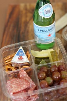 a plastic container filled with meat, cheese and grapes next to a bottle of beer