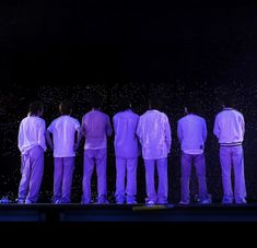 five men in white shirts and purple pants stand on stage with their backs to the camera