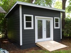 a small gray shed with white trim on the windows and door is next to a tree