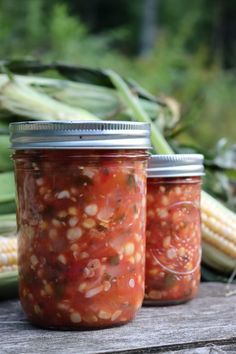 three jars filled with pickled vegetables sitting on top of a wooden table next to corn