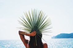 a woman standing on the beach with her back to the camera holding a palm leaf over her head