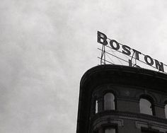 a black and white photo of the boston sign on top of a tall building in front of a cloudy sky