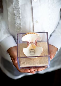 a person holding a ring in a glass box with sand and sea shells on it