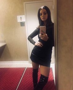 a woman standing in front of a mirror looking at her cell phone while wearing knee high boots