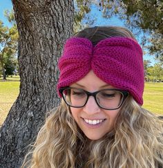 a woman wearing glasses and a pink knitted headband smiles at the camera while standing in front of a tree