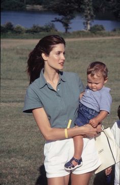 a woman holding a small child in her arms while standing next to another person on the grass