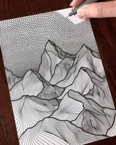 a person is drawing on paper with a pen and inking it in the shape of mountains