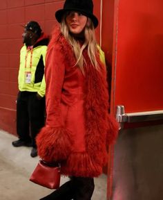 a woman in a red coat and black hat is standing by a wall with her hands on her hips