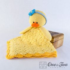 a crocheted yellow ducky with a blue bow on its head sits next to a piece of wood