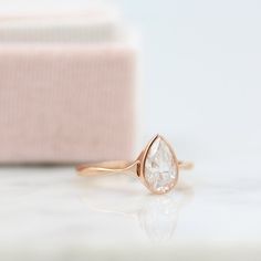 a gold ring with a pear shaped diamond in it on a marble surface next to a pink box