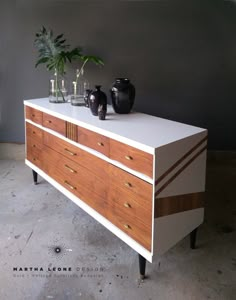 a white and brown dresser with two vases on it's top, next to a potted plant