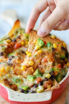 a hand dipping a tortilla chip into a bowl full of mexican food with corn and avocado