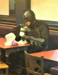 a person in a batman costume sitting at a table drinking from a cup with the caption, i'll have a 7