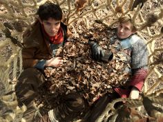 two people are sitting on the ground surrounded by dead leaves and branches, looking up at something in the air