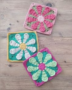 three crocheted squares with flowers on them sitting on a wooden floor, one is pink and the other is green