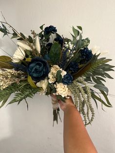 a person holding a bouquet of flowers and greenery in their hand, against a white wall