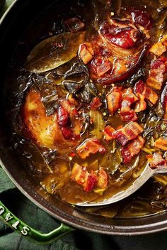 a pan filled with meat and vegetables on top of a green table cloth next to a wooden spoon