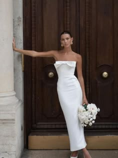 a woman standing in front of a brown door wearing a white dress and holding her arm out