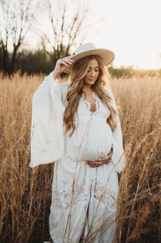 a pregnant woman standing in tall grass wearing a white dress and hat with her hands on her head