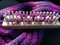 a purple knitted scarf with pins stuck in the middle on a wooden holder for knitting needles