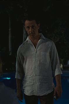 a man standing in front of a pool at night wearing a white shirt and black pants