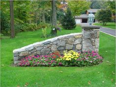 a stone wall and flower bed in the middle of a lawn with trees behind it