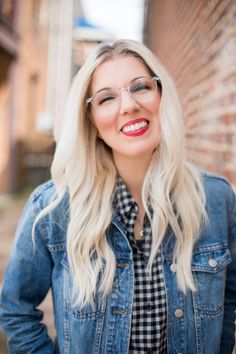 a woman with blonde hair and glasses standing in front of a brick wall smiling at the camera