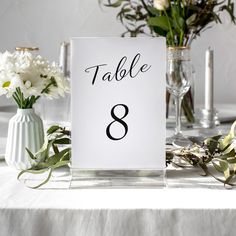 a table number with flowers and vases in the background on a white table cloth