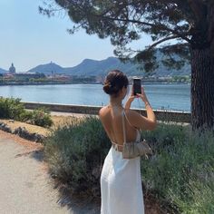 a woman in a white dress taking a photo with her cell phone near the water