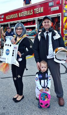 two adults and a child standing in front of a firetruck with costumes on