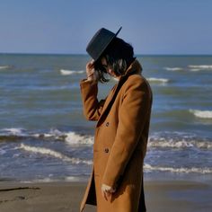 a person standing on the beach wearing a coat and hat with one hand to his ear