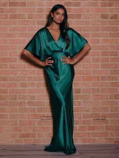 a woman standing in front of a brick wall wearing a long green dress with cape sleeves