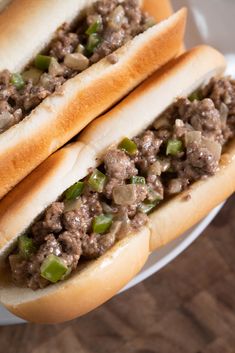 two hotdogs with meat and green peppers on a white plate sitting on a wooden table