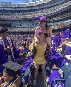 two women in graduation gowns sitting on the stands at a stadium with other people