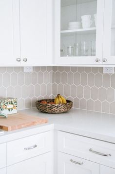 a kitchen with white cabinets and wooden cutting board on the counter top next to a basket filled with fruit