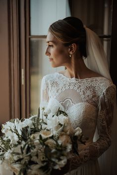 bride in a long sleeve lace wedding dress looking out the window holding a white and green wedding bouquet Lace Mock Neck Wedding Dress, Mock Neck Long Sleeve Wedding Dress, Maid Of Honour Speech, White And Green Wedding Bouquet, Elegant Lace Wedding Dress, Bride Surrounded By Bouquets, Wedding Flowers White, Long Sleeve Lace Wedding Dress, Sleeve Lace Wedding Dress