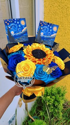 a hand holding a bouquet of blue and yellow flowers with pictures on the top, in front of a window