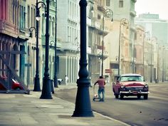 an old red car driving down a street next to tall buildings on either side of a lamp post